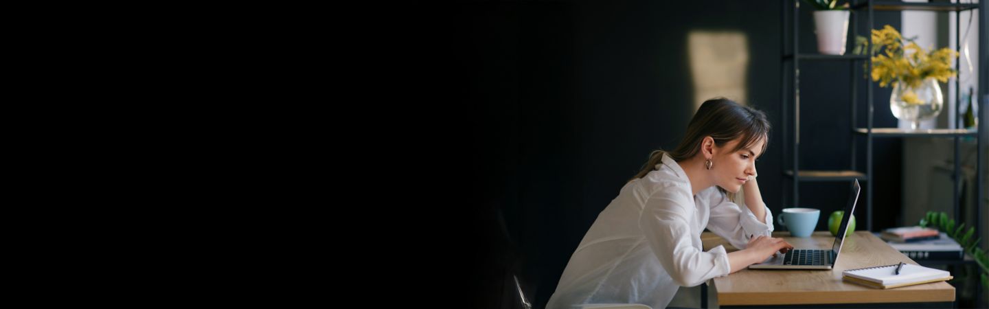 Млада жена седи на биро и концентрирано работи на лаптоп, со тетратка и шолја до неа. Позадината е темна, а фокусот е на младата жена.