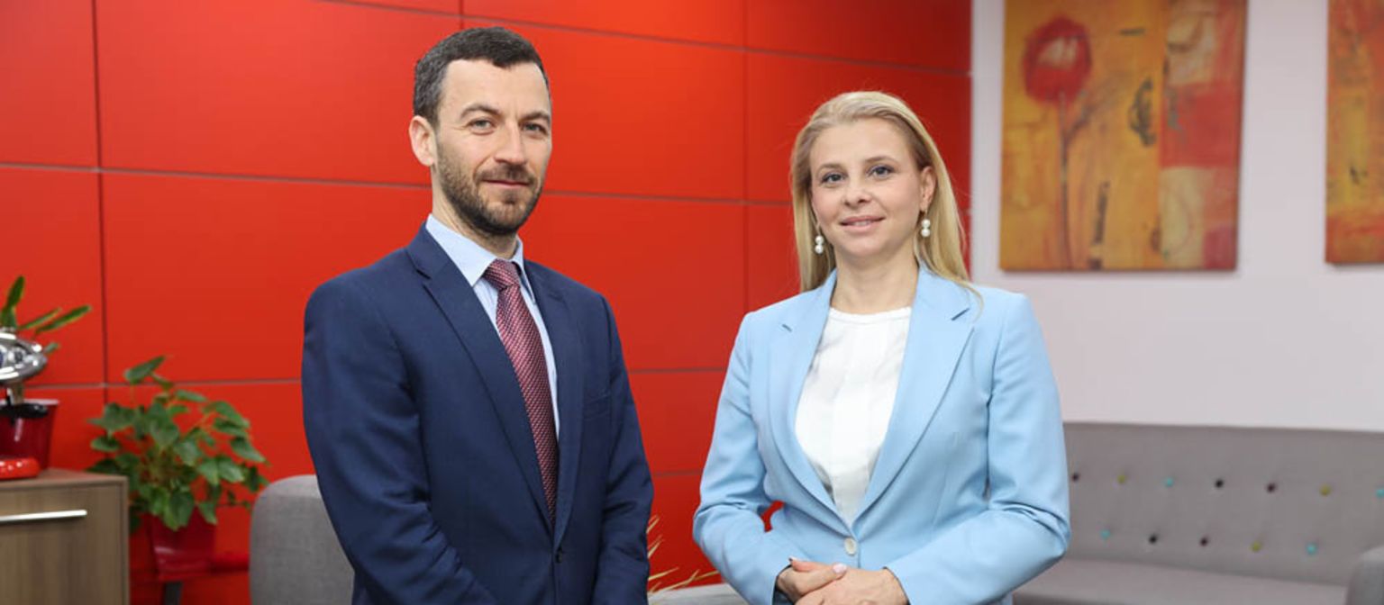 Извршниот директор Владислав Хаџидинев во сино одело и катерина Бошевска извршен директор на ЕОС Северна Македонија во светло сино блејзер кој стои еден до друг во канцеларија со црвен ѕид