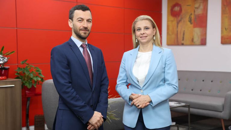 Извршниот директор Владислав Хаџидинев во сино одело и катерина Бошевска извршен директор на ЕОС Северна Македонија во светло сино блејзер кој стои еден до друг во канцеларија со црвен ѕид