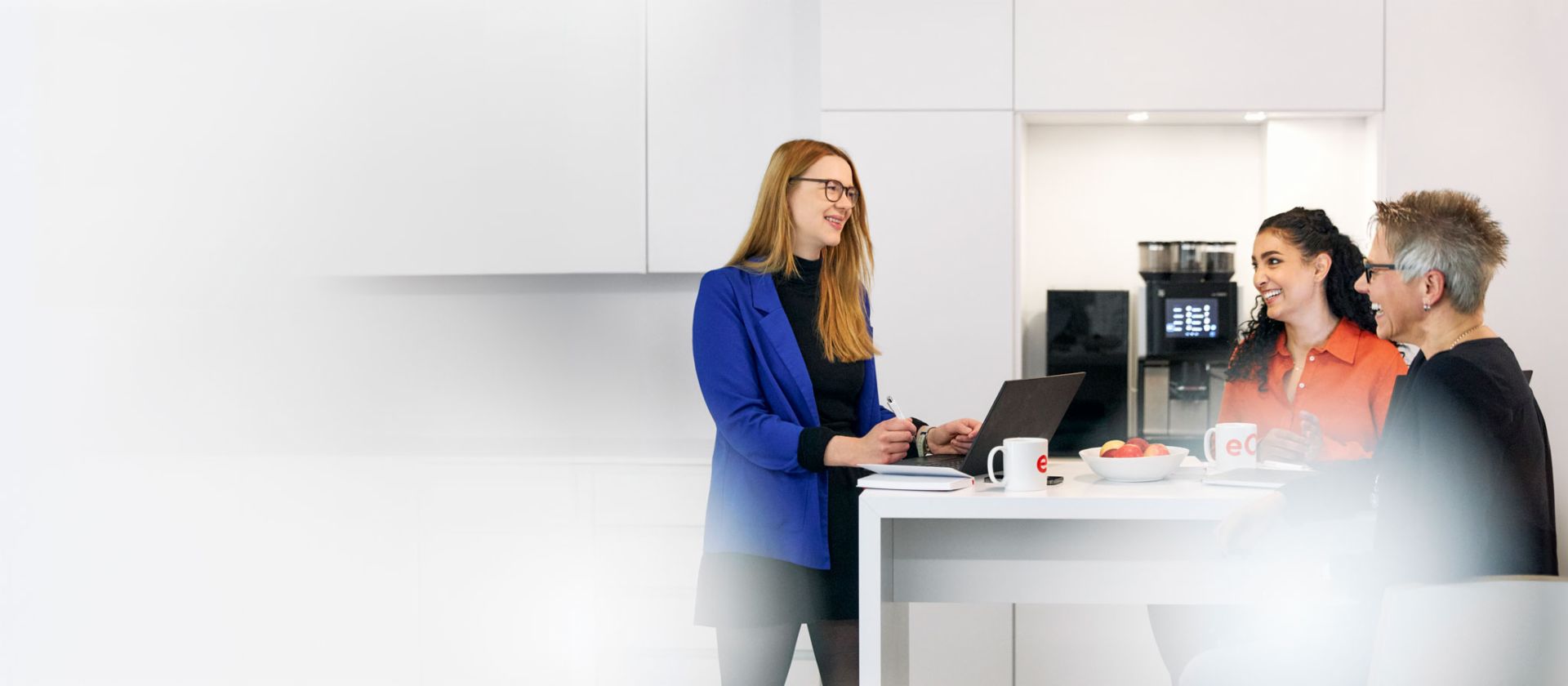  Три лица во модерен канцеларија работат на маса со EOS шолји и лаптоп, додека четврта личност стои на кафематот во позадина - Тимска работа и кариерни можности во EOS