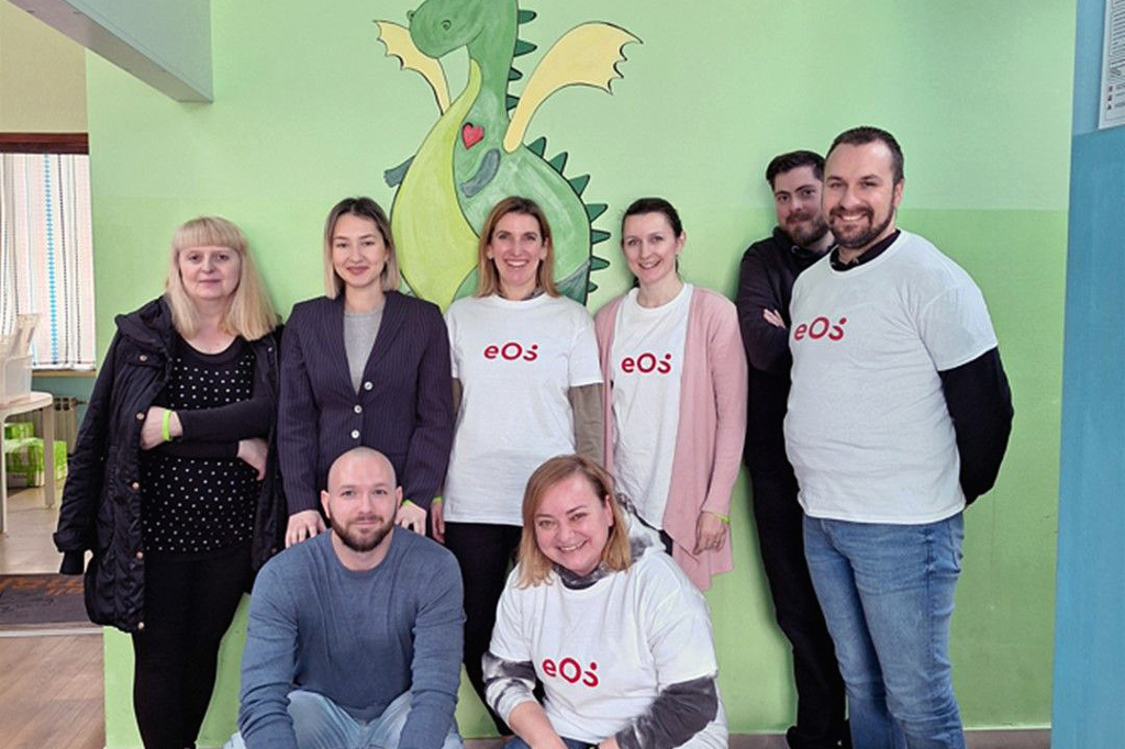 Осум луѓе стојат пред ѕид на кој е насликан зелен змеј. Луѓето се наредени во два реда: двајца се клекнати напред, а шестмина стојат одзади. Четири лица носат бели маици со црвеното лого на EOS. Групата се насмевнува пред камерата. На подот, напред десно, гледаме неколку шишиња и кутии за пијалаци.