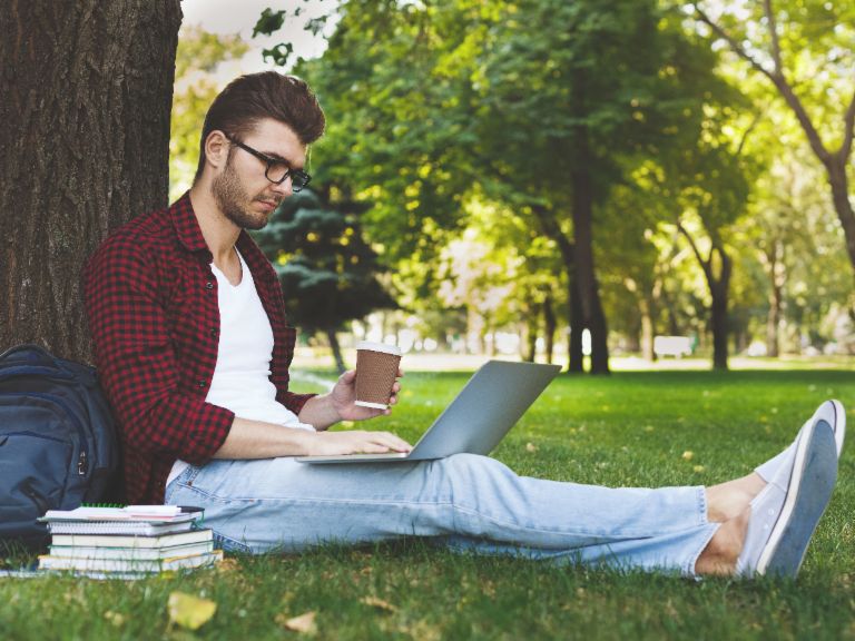 Image of a student sitting under a tree with their laptop, books and a coffee cup.