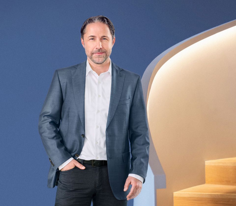 Марвин Рамке, Главен извршен директор на EOS Group, стои пред сина позадина, облечен во темно сако. Тој гледа директно во камерата.
