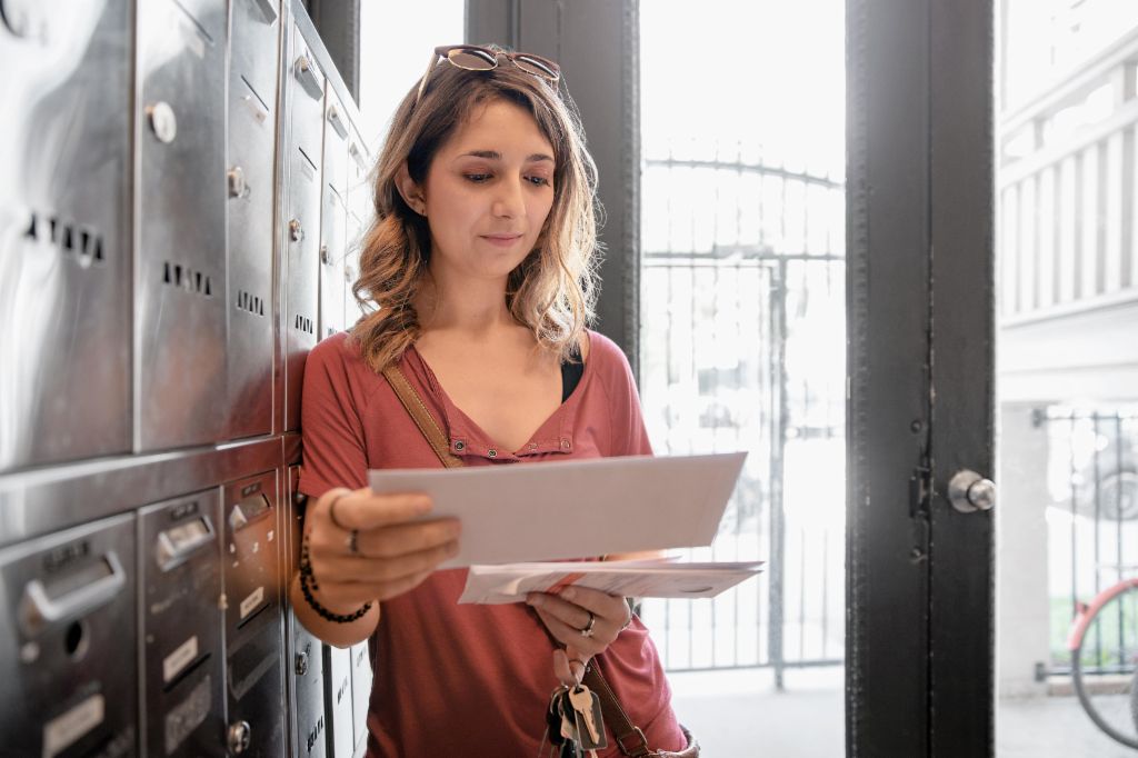 Жена со руса коса се потпира на редица поштенски кутии, размислувајќи додека го проверува писмото што токму го добила.